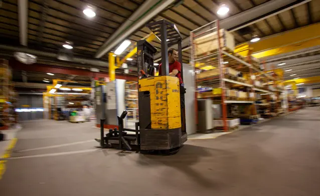 A forklift in a large warehouse with lights on.