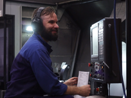 En man i blå skjorta och hörselskydd arbetar vid en maskin.