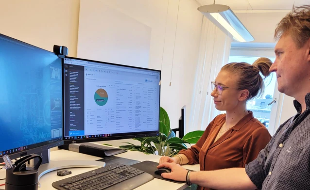 Two people are standing in front of a computer, where one is demonstrating and explaining how the Monitor ERP system works to the other.