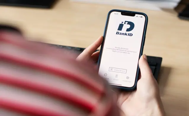 A person holding a cell phone over a desk with BankID visible on the screen.